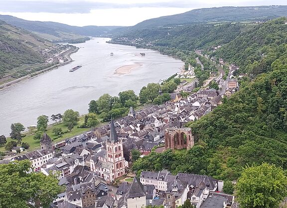 Das Bild zeigt einen malerischen Blick auf Bacharach, eine Stadt am Rhein. Im Vordergrund befindet sich eine Ansammlung von Gebäuden, darunter eine markante Kirche mit hohem Turm. Die Stadt liegt eingebettet zwischen üppig grünen Hügeln, die auf beiden Seiten steil ansteigen. Rechts ist auf einem Hügel eine große Burg zu sehen, umgeben von dichtem Wald. Ein weiterer kleinerer Turm befindet sich weiter unten am Hügel. Der Rhein fließt entlang der Stadt, mit einer sichtbaren Sandbank in der Mitte des Flusses. Der Fluss schlängelt sich sanft ab und erstreckt sich in die Ferne, umgeben von weiteren Hügeln und Grün.