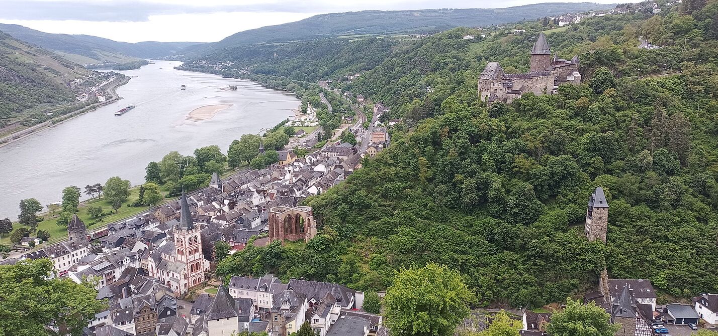 Das Bild zeigt einen malerischen Blick auf Bacharach, eine Stadt am Rhein. Im Vordergrund befindet sich eine Ansammlung von Gebäuden, darunter eine markante Kirche mit hohem Turm. Die Stadt liegt eingebettet zwischen üppig grünen Hügeln, die auf beiden Seiten steil ansteigen. Rechts ist auf einem Hügel eine große Burg zu sehen, umgeben von dichtem Wald. Ein weiterer kleinerer Turm befindet sich weiter unten am Hügel. Der Rhein fließt entlang der Stadt, mit einer sichtbaren Sandbank in der Mitte des Flusses. Der Fluss schlängelt sich sanft ab und erstreckt sich in die Ferne, umgeben von weiteren Hügeln und Grün.